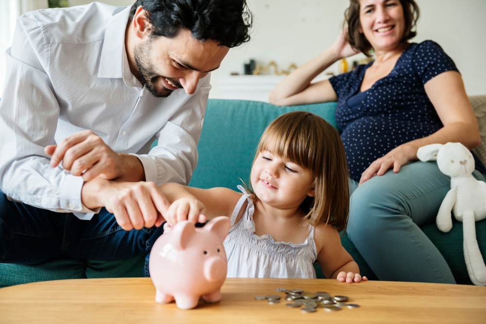 Parents watching little girl put coins in piggy bank